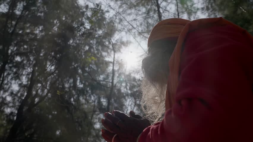 An elderly Indian sadhu dressed in saffron robes sits cross-legged in a serene forest setting, blowing a traditional horn during a spiritual ritual. With a long white beard, turban, and sacred beads around his neck, he appears deeply focused and immersed in meditation. Natural sunlight filters through the trees, creating a peaceful and spiritual atmosphere.