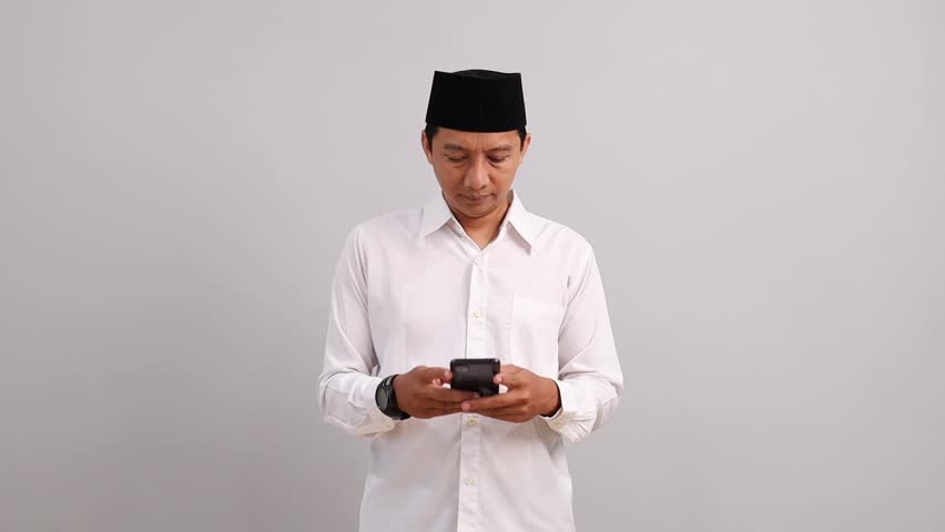 Asian Muslim man using his mobile phone, looking sideways, surprised to see something next to him in the empty space while smiling.