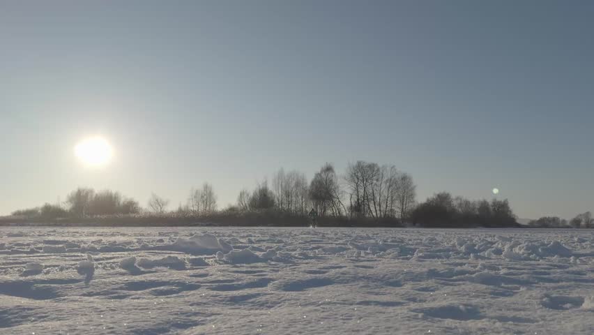 Motocross rider performing a wheelie on a frozen lake while moving toward the camera. Dirt bike with studded tires riding on ice under clear winter sky, minimal snowy landscape with no buildings or infrastructure in the background.