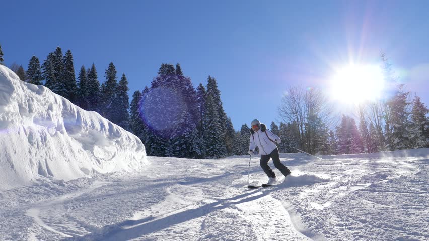 Adult skier sliding down a mountain slope covered with fresh powder snow on beautiful sunny winter day with blue sky. Sun rays shines ski piste surrounded by coniferous forest in ski resort