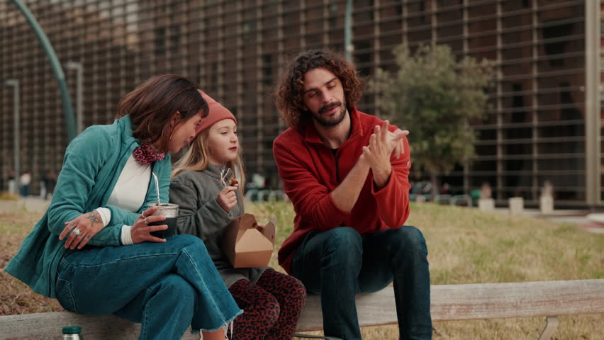 A happy family, including a mother, father, and daughter, shares a moment of connection outdoors. They are seen laughing and talking while enjoying snacks and drinks on a sunny day.