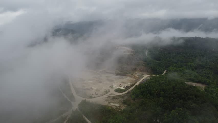 A mist-covered open quarry in Trbovlje, Slovenia, seen from above, with winding dirt roads, exposed earth, and dense green forested hills fading into low clouds.