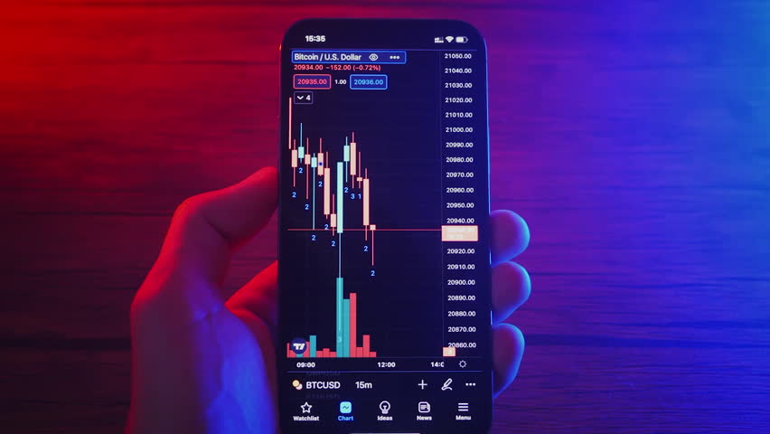 A close-up view of a hand holding a smartphone that shows a Bitcoin price chart with colorful candlestick indicators. The background features a gradient of red and blue lighting.