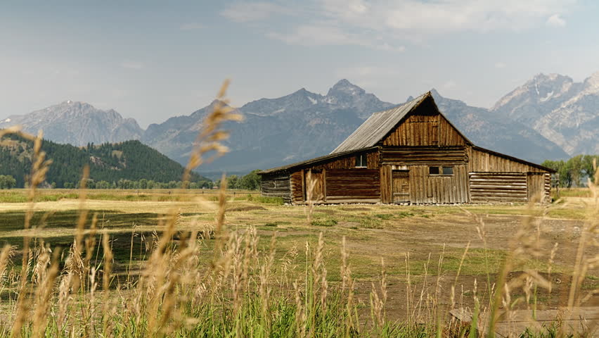 A weathered barn sits in a valley with the Teton Mountain Range in the background; peaceful, static shot.