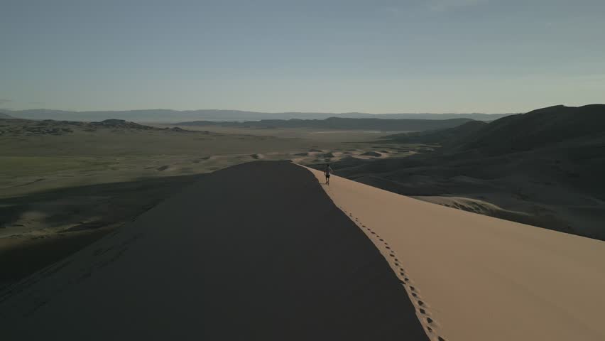 Aerial view of traveler walking on top of sand dune at sunset, drone follow shot from behind, footprints on sand ridge, vast steppe panorama in Mongolia, golden hour, 4k raw unedited footage