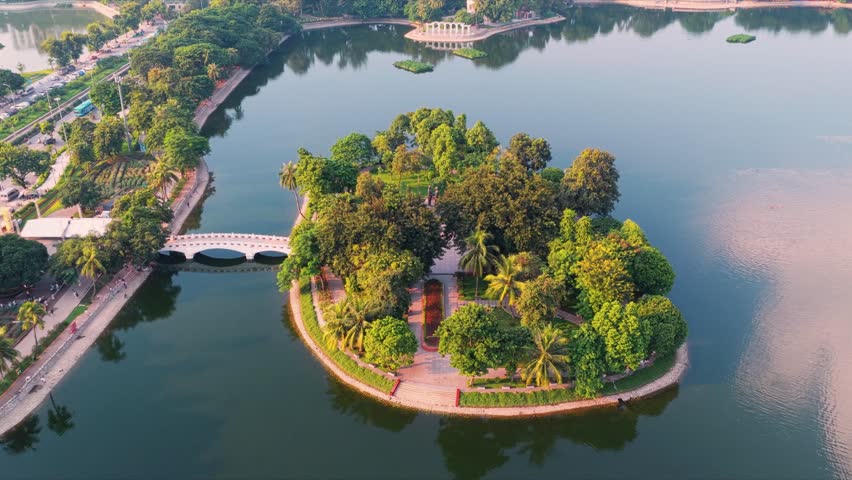 Autumn sunset over Thong Nhat Park in Hanoi, Vietnam, aerial view. This high-angle drone shot captures the golden sunlight filtering through yellow-leaf trees and shimmering on the central lake during a peaceful autumn afternoon, highlighting the lush green landscape and urban park scenery.