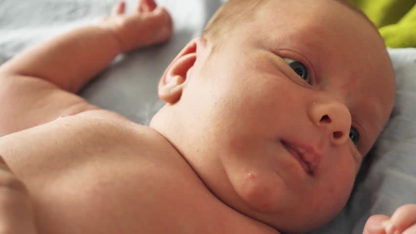 Close-up of an alert three-week-old infant lying on a soft blanket. The baby has blue eyes, open and curious, and makes a funny face sticking its tongue out. Soft, natural lighting