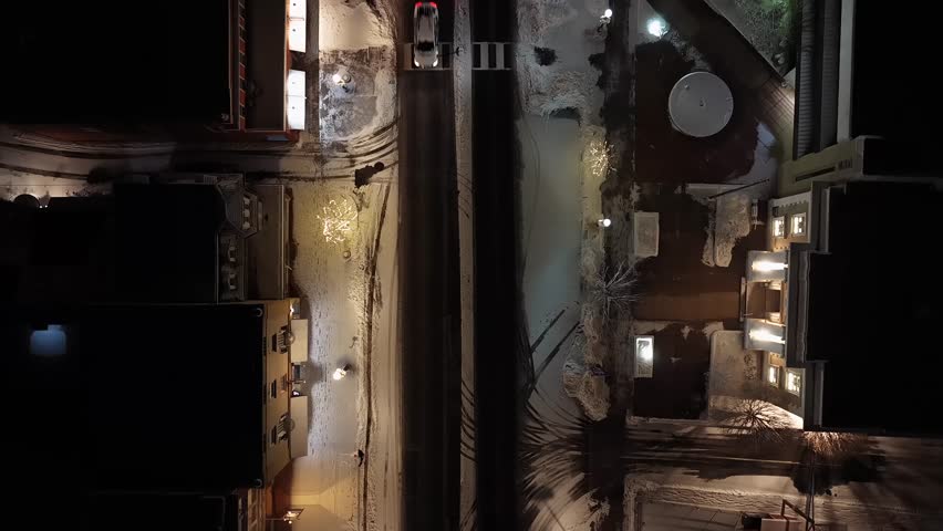 Quiet American street at night, single car driving through snow lined roads, warm street lights illuminating winter sidewalks and residential buildings. Aerial top down shot. Christmas time in USA.