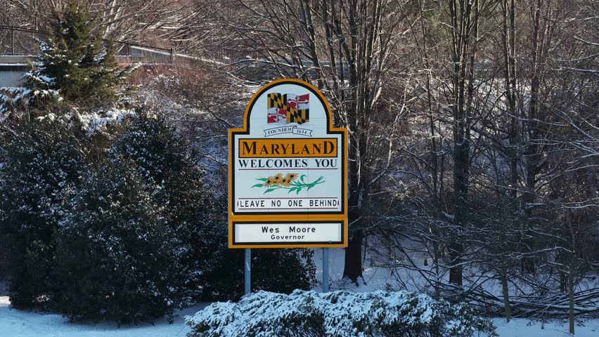 Aerial zoom shot of Maryland welcome sign surrounded by snow-covered trees, at quiet winter border scene with fresh snowfall and calm seasonal atmosphere. Pennsylvania and Maryland border in USA.