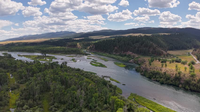 Drone footage of the Snake River winding through a green valley with forested banks and distant hills under a partly cloudy sky.
