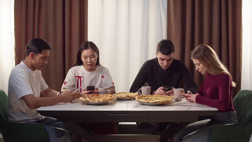 Group young adult friends with cell mobile phone addiction having lunch together. People enjoy pizza drink tea at home. Awkward ignore gathering problem. Students team meeting scroll. Modern youth.