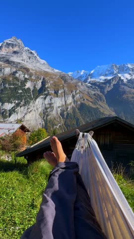 Relaxing swing bare feet Hammock Murren Mürren Gimmelwald Lauterbrunnen Valley sunny autumn fall blue sky Swiss Alps Switzerland Schweiz vertical reel grass cliff slabs Allemdhubel Jungfrau mountain