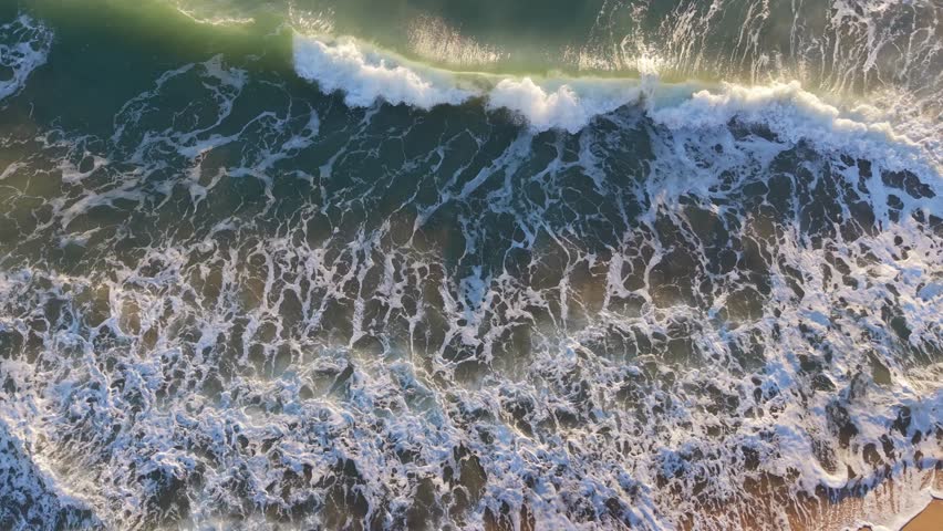 Beautiful coastal seascape filmed from above with turquoise water and crashing waves forming textures beside sandy shoreline.