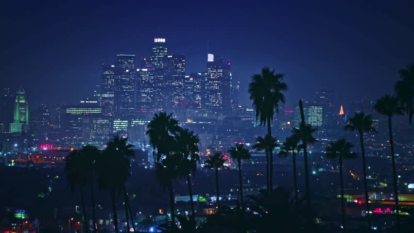 Dazzling night view of illuminated Downtown Los Angeles skyscrapers framed by silhouettes of iconic palm trees.