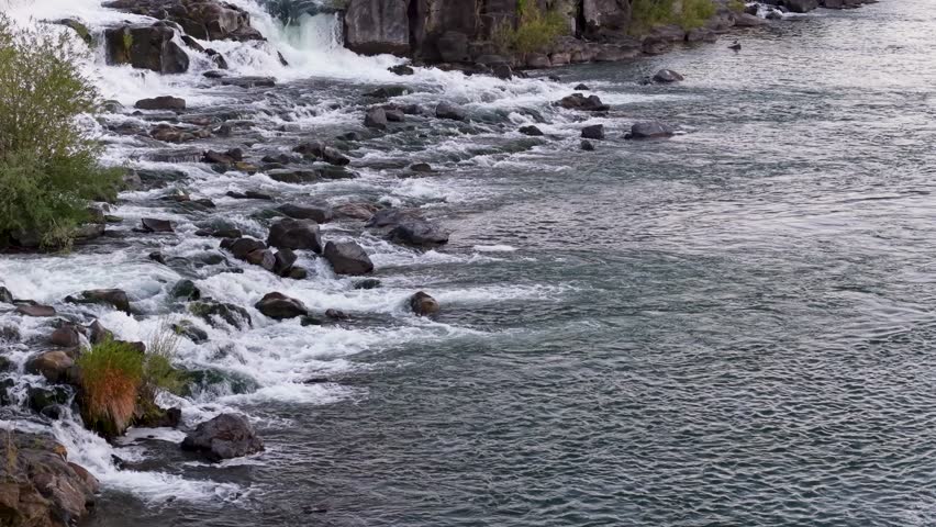 An aerial view of a rocky river where rushing water tumbles over stones, creating small rapids and swirling currents Idaho Falls