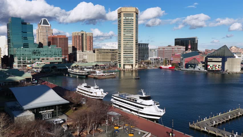 Aerial view of Baltimore Inner Harbor on sunny winter day with boats and ships docked along waterfront, calm water and clear city skyline in background. Rising shot. Traffic on Main Street.