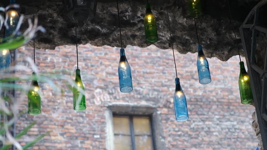 Hanging glass bottle lights suspended on cables against a brick wall background in a courtyard.