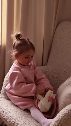 A vertical shot of a toddler girl in a pink dress sitting in a chair and looking at her reflection in a white heart-shaped toy mirror. Concept of childhood vanity and play.