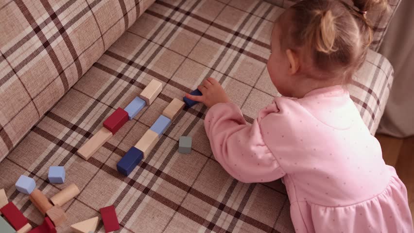 A high-angle shot of a little girl in a pink dress carefully arranging colorful wooden blocks in a row on a checkered sofa. focus on fine motor skills.