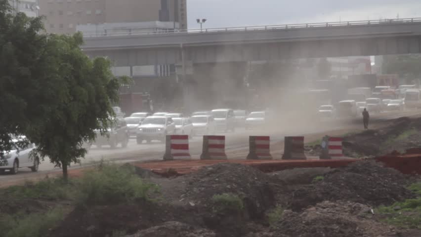 Traffic crawls past dusty roadworks beneath an elevated overpass in Nairobi, Kenya, with construction barriers, earth piles and haze in afternoon light—urban infrastructure scene