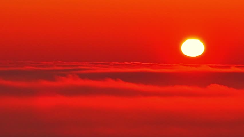 Drone view of the sun above a sea of clouds at dusk. Deep red tones fill the sky as sunlight fades along the horizon, creating a dramatic, abstract background with strong atmospheric mood.