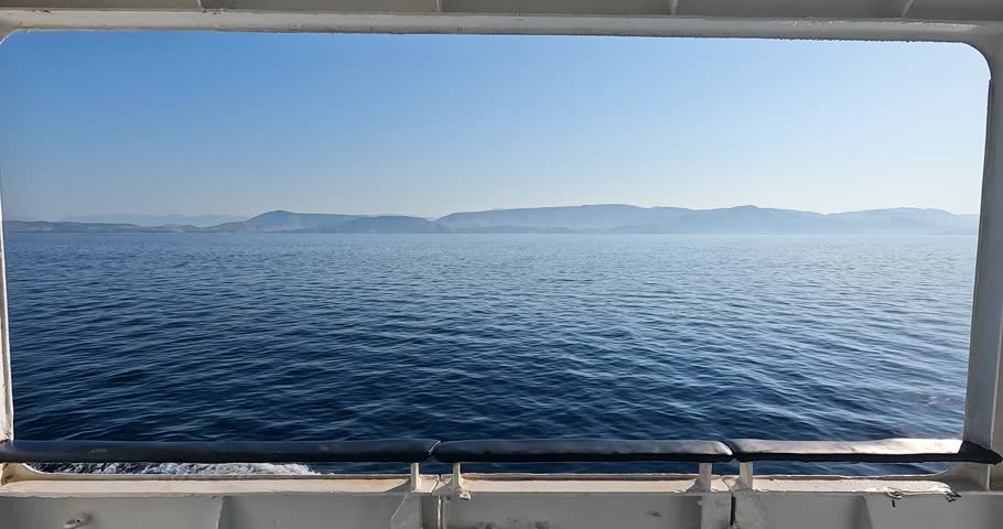 Scenic view of the deep blue Ionian Sea and distant hazy mountains framed by a large ferry window.