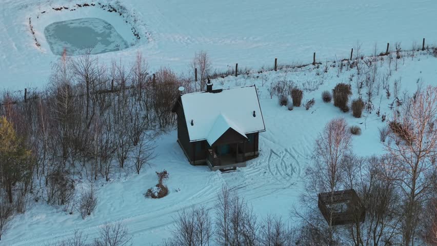 Aerial view of a remote wooden cabin surrounded by snowy fields and bare trees, capturing winter solitude and rural isolation.