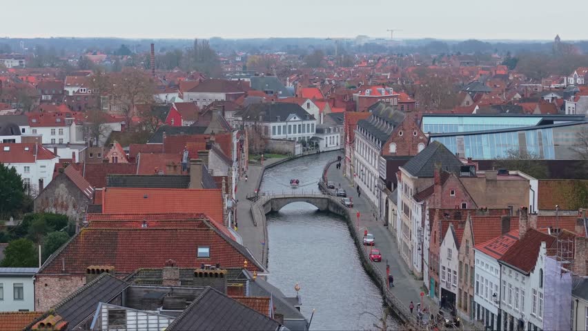 Drone footage of the historic canals in Bruges, Belgium, during a moody winter day. Overcast sky reflecting on the water surrounded by traditional Flemish architecture and ancient brick houses.