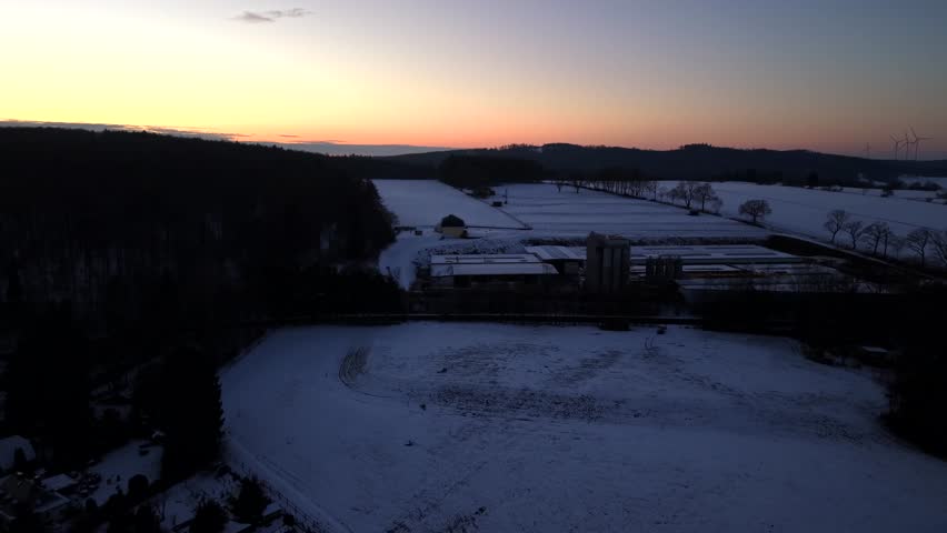 Aerial view of American industrial area at winter sunset, showing snow-covered fields and factory warehouses with warm evening light on horizon and calm rural landscape. Wide shot.