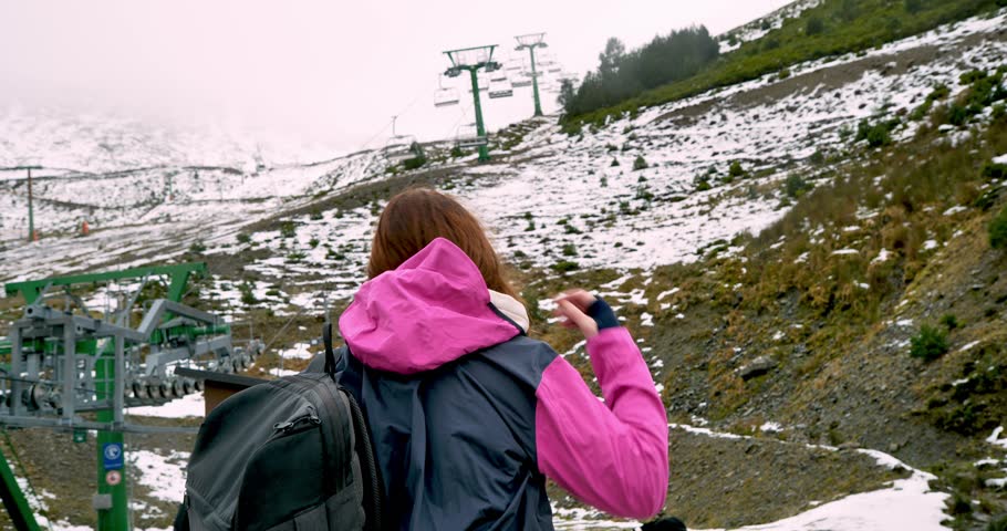 On a winter day, a woman puts on the hood of her technical jacket while watching the ski lift chairs.