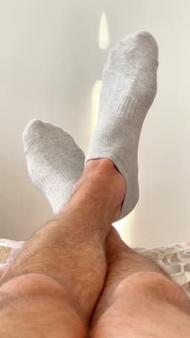Close-up of a man's legs and feet wearing grey socks, casually crossed and moving slightly while he is relaxing in bed on a sunny morning, conveying a sense of comfort and leisure at home