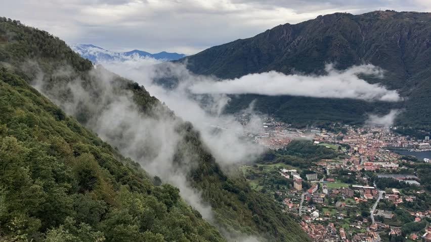 Slow motion clouds drifting over a mountain valley and small town below, atmospheric landscape with mist and fog creating a calm and cinematic nature scene in HD.