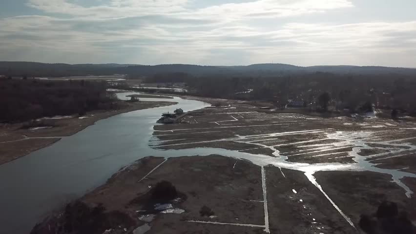 Drone aerial of the Essex River winding through the Great Marsh in winter, with reflective tidal channels, salt marsh flats, and a small building on the riverbank, Massachusetts.