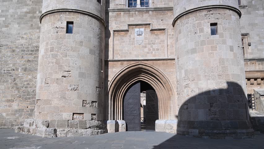 Slow upward pan revealing the fortified stone walls and towers at the entrance of the Palace of the Grand Master of the Knights of Rhodes in the town of Rhodes, Greece under clear blue sky
