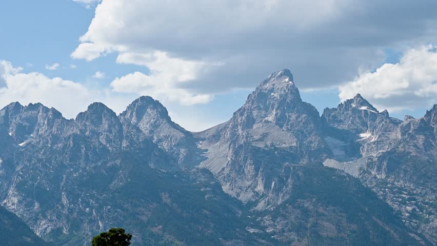A dramatic mountain range with rugged peaks rises sharply under a partly cloudy blue sky, capturing the majestic beauty of the landscape, Grand Teton National Park