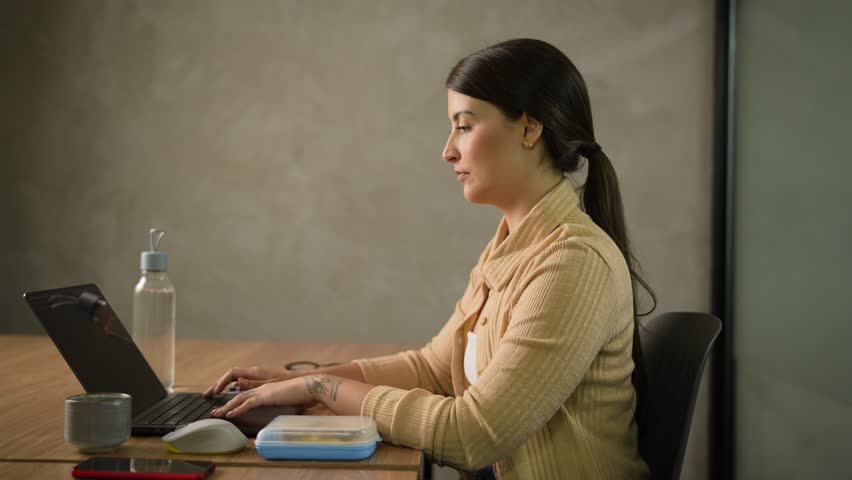Female freelancer with arm tattoo eating homemade sandwich during lunch break at wooden desk in contemporary office space