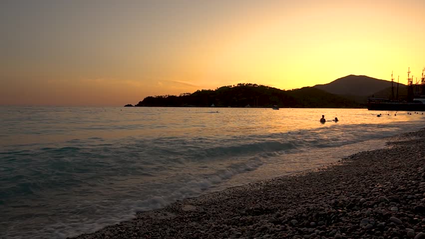 The setting sun casts golden light on the seashore of a tropical resort. Silhouettes of people swimming are visible in the ocean waves.