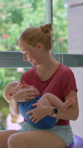 Young mother sits on a fit ball gently rocking her newborn son in her arms by a large window with lush tropical greenery, helping him fall asleep with soothing motion and care.