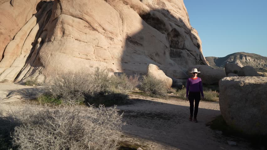 A woman walks along a hiking trail in Joshua Tree National Park surrounded by rocks and desert plants as the sun sets in the background.