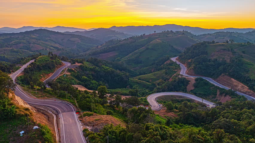 Aerial view of Golden sunset light glowing over a winding mountain road on a high ridge in Nan Province, Thailand, creating a dramatic landscape of curves and towering peaks.