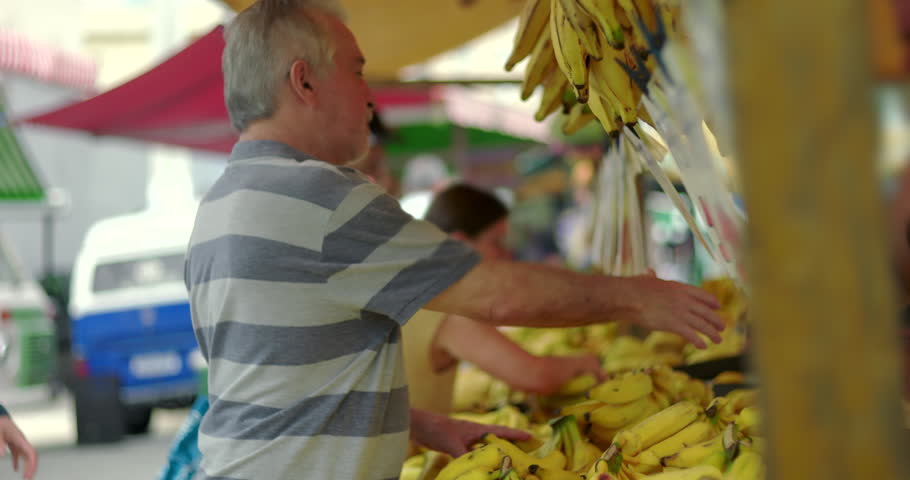 Senior shopper welcoming vendor before checking bananas for purchase conveying neighborly exchange familiarity and natural participation in community street market life