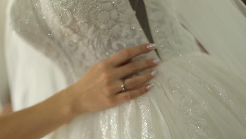 A bride in a white satin wedding dress with pearl accents holds a delicate bouquet of white flowers and baby