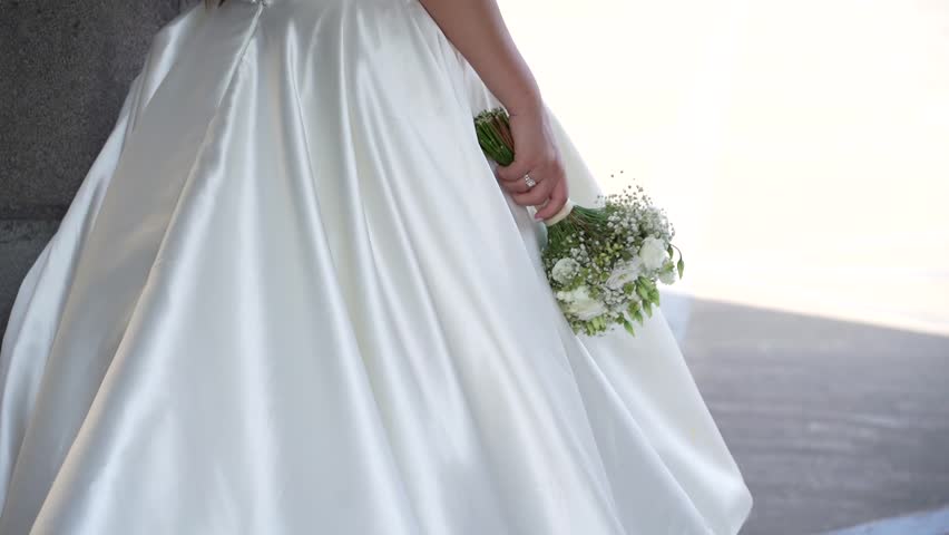 A bride in a white satin wedding dress with pearl accents holds a delicate bouquet of white flowers and baby