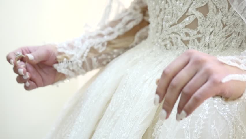 A bride in a white satin wedding dress with pearl accents holds a delicate bouquet of white flowers and baby's breath, showcasing her ring.