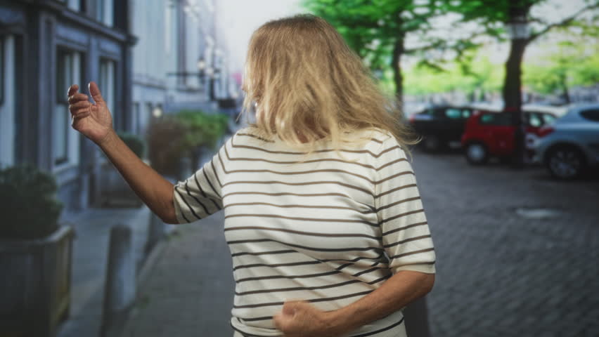 Woman with blonde hair covering face turning and dancing with arm extended on a street; carefree joy.
