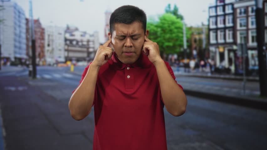 Young hispanic man in red polo shirt with hands covering ears on a busy city street; sensory overload stress.