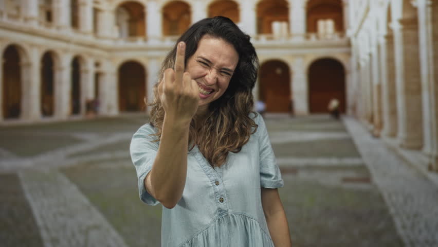 Woman showing middle finger in a sunlit building courtyard with arched corridors and worn stone floors; defiance.