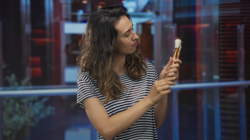 Brunette woman wearing a striped shirt carefully examining makeup brushes in a hotel interior; curiosity.