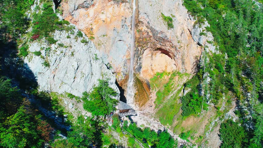 Aerial view from above of a waterfall stream descending along a steep rock face. Clear mountain water flows through rugged terrain in summer daylight.
