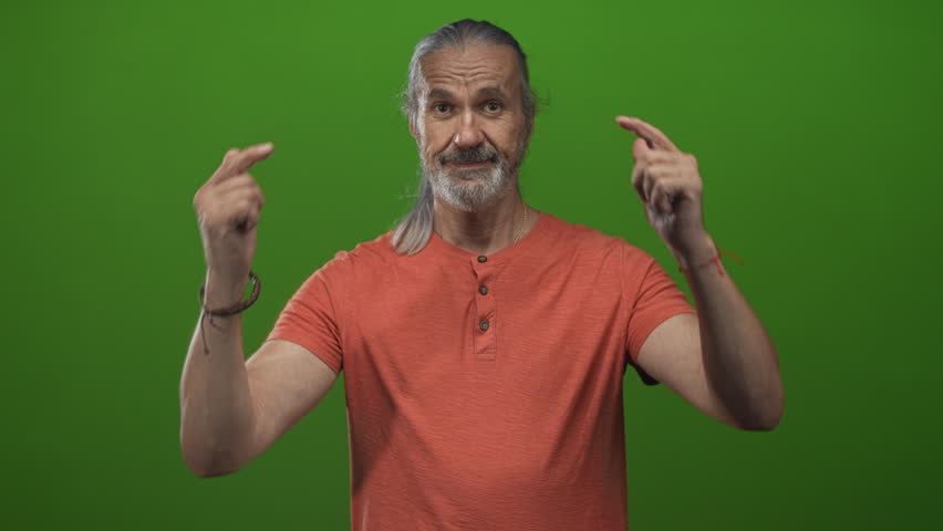 Man points both fingers down in a green studio, hoary long hair and beard, wearing orange shirt; self promotion playful.